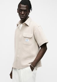 Άνδρας με μπεζ κοντομάνικο υφασμάτινο πουκάμισο με τσέπη στο στήθος και λευκό παντελόνι, στέκεται με το ένα χέρι στην τσέπη, μπροστά σε λευκό φόντο.