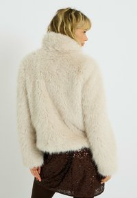 Veste en fausse fourrure beige clair, design court avec un col montant, texture pelucheuse et manches larges, associée à une jupe brune pailletée.