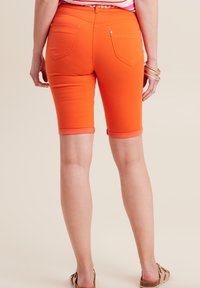 Shorts bermuda orange en tissu extensible, avec des ourlets roulés et des poches arrière. Portés avec un haut rayé décontracté et des sandales.