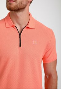 Lühikeste varrukatega polo särk erksas korallis, millel on tekstuuriga muster, pooldatav krae ja väike logo rinnal.
