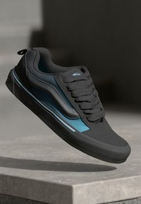 Zwarte sneaker met een gladde textuur, voorzien van een blauwe zijpaneel, zwarte veters en een laag profiel ontwerp. Subtiele accenten en logo zichtbaar.