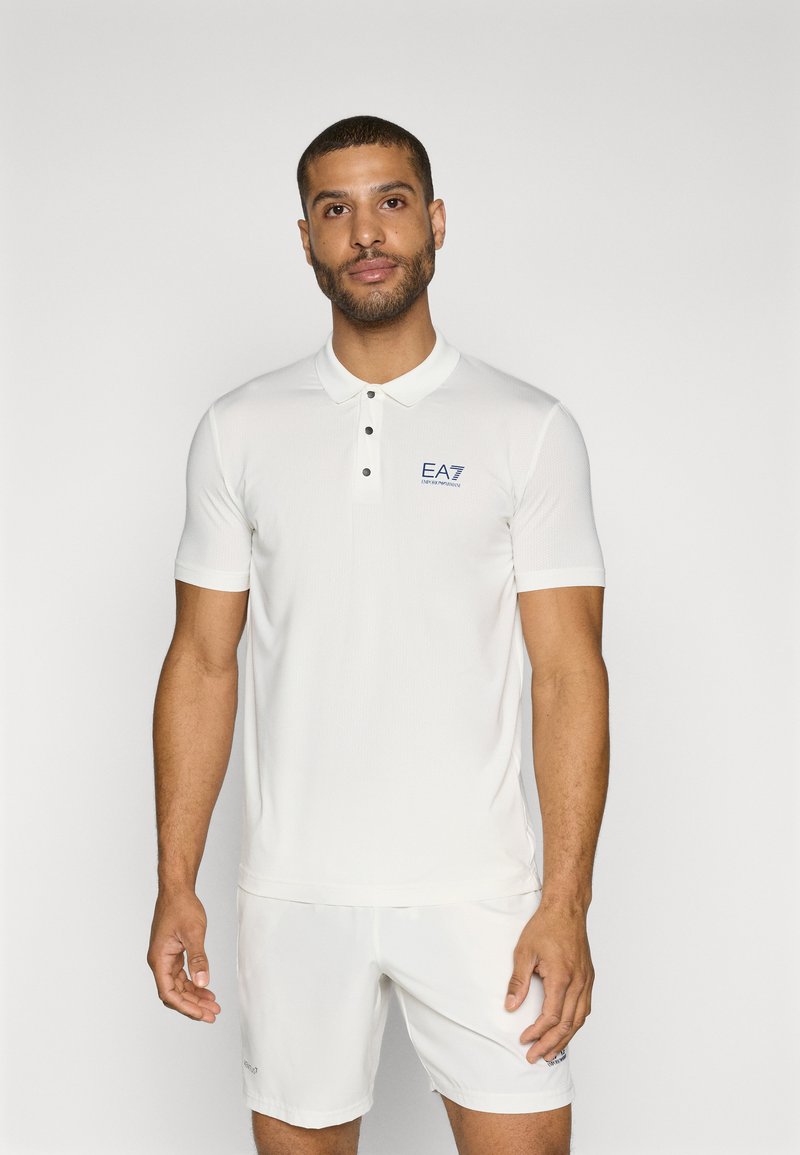 Λευκό κοντομάνικο πουκάμισο πόλο με υφασμάτινη υφή, με πλεχτό κλείσιμο δύο κουμπιών και μπλε λογότυπο EA7 στο αριστερό στήθος. Φορεμένο με λευκές βερμούδες.