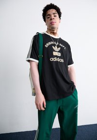 Melna kokvilnas T-krekls ar baltu apmali un zeltītu Adidas logo, savienots ar zaļām sporta biksēm, kurām ir baltas sānu svītras un izšuvuma detaļas.