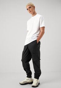 T-shirt bianca in cotone, pantaloni cargo neri con tasche con zip e sneakers beige con un design ondulato nero. Vestibilità casual, texture liscia.