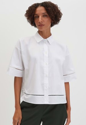 Vrouw met kort krullend haar draagt een wit, kort mouwig, knoophemd met horizontale uitsnijdingen aan mouwen en zoom, zwarte broek.