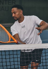 Camiseta deportiva blanca con logo negro, combinada con pantalones cortos negros. El jugador sostiene una raqueta de tenis naranja cerca de una red en una pista azul.
