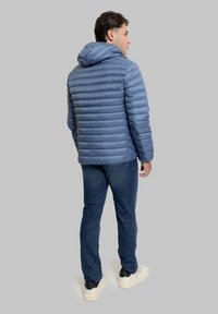 Giubbotto imbottito blu con design trapuntato orizzontale, cappuccio e cerniera frontale. Indossato sopra jeans scuri in denim e scarpe da ginnastica chiare.