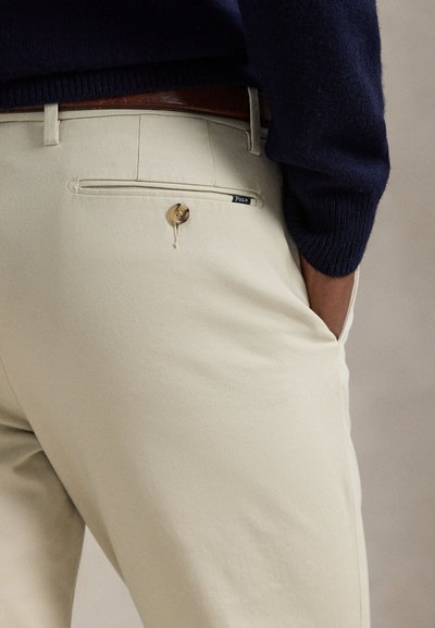 Osoba nosiaca béžové nohavice s zapnutým zadným vreckom na gombík a hnedým opaskom, ruka v prednom vrecku, vidieť tmavomodrý sveter.