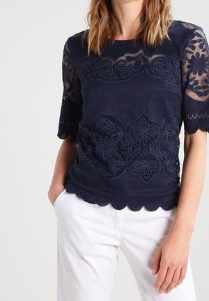 Femme portant un haut en dentelle bleu marine avec des motifs floraux et des bords festonnés, associée à un pantalon blanc, debout avec une main dans la poche.