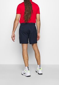 Pantalones cortos navy con cintura elástica, camiseta polo roja y zapatillas blancas con acentos neón. Diseño limpio y sencillo, apropiado para un look casual.