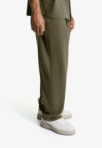 Pantaloni de culoare verde măsliniu, confecționați dintr-un material neted, cu un croi lejer. Asortați cu adidași albi cu accente gri și tălpi texturate.