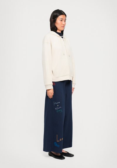 Woman wearing cream zip-up hoodie and navy wide-leg pants with colorful text "Have a Nice Day" and "Love," standing in profile.