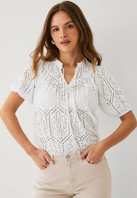 Hvid bluse med ørekant og V-udskæring, korte ærmer og intrikate udskæringsmønstre. Parret med lyse beige bukser, der har en afslappet pasform.