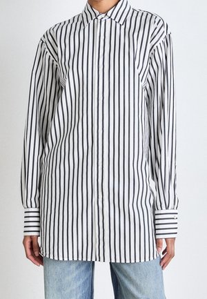 Hvid langærmet skjorte med lodrette sorte striber iført over lyseblå jeans, vist fra nakke til hofter.