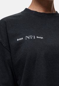 Zwarte katoenen sweatshirt met een ronde hals, voorzien van witte geborduurde tekst die luidt: "British No1 Design." De textuur is zacht en mat.