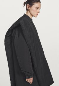 Cappotto oversize nero con texture trapuntata. Presenta un colletto alto e maniche larghe, creando una silhouette a forma di bozzolo.