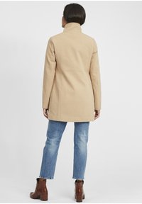 Beige Wollmischmantel mit hohem Kragen, taillierter Silhouette und mittlerer Länge, kombiniert mit hellblauen Jeans und braunen Stiefeletten.