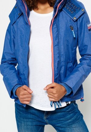 Osoba w niebieskiej kurtce na zamek z koralową wyściółką zamka, białą koszulą i niebieskich dżinsach, trzymająca krawędzie kurtki obiema rękami.