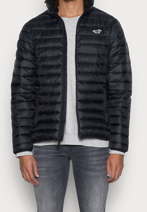 Schwarze Daunenjacke mit Reißverschluss vorne, horizontal gestepptem Design und einem kleinen weißen Logo auf der oberen linken Brust. Trägt man über einem grauen Shirt.