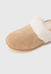 Beige semsket slippers med fluffy hvit lammeskinnforing og en sydd såle kant, vist i nærbilde mot en hvit bakgrunn.