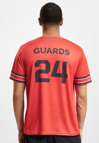 Rød sportsdrakt med "GUARDS" og "24" i fet svart skrift. Korte ermer med svart og hvit stripete mansjetter. Pustende stoff.