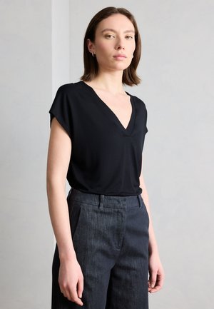 Young woman with shoulder-length brown hair wearing a black V-neck blouse and dark gray pants against a light gray background.