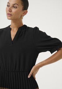 Schwarzes Kleid mit kurzen Puffärmeln, einem V-Ausschnitt und einem gefältelten unteren Teil. Glatter Stoff mit einem figurbetonten Oberteil, das in eine fließende Textur übergeht.