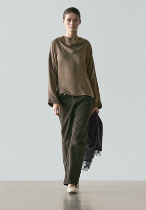 Donna che indossa una maglia marrone larga a maniche lunghe, pantaloni larghi scuri, ballerine bianche, che tiene una sciarpa scura con frange mentre cammina su un pavimento grigio.