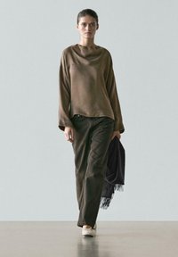 Vrouw draagt loszittende bruine lange mouwen top, donkere broek met wijde pijpen, witte ballerina's, houdt een donkergrijze sjaal met franjes vast terwijl ze over een grijze vloer loopt.