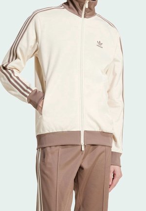 Mężczyzna ubrany w beżową i brązową dresową kurtkę i spodnie Adidas z trzema paskami na rękawach i bokach spodni.