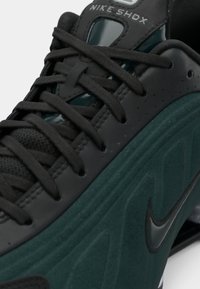 Sapatilhas Nike Shox pretas e verde-escuro com atacadores pretos e logotipo da Nike em destaque de lado.
