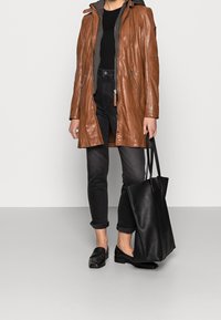 Bruine leren jas met ritssluitingen, grijze capuchon en gestructureerde afwerking. Gecombineerd met zwarte jeans, zwarte loafers en een grote zwarte schoudertas.