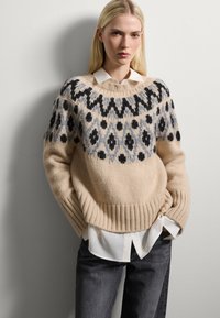 Frau mit langen blonden Haaren, die einen gemusterten beigen Strickpullover über einem weißen Hemd und dunklen Jeans trägt, steht vor einer schlichten Wand.