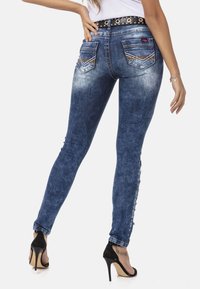 Skinny jeans in middelblauw denim, met een versleten textuur, achterzakken met chevron-stiksel en een zwarte riem met metalen accenten.