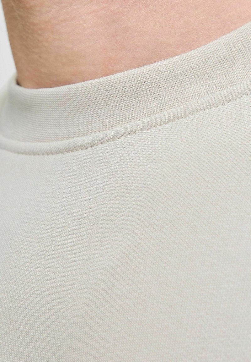 Gros plan sur le cou d'une personne portant un t-shirt beige clair à col rond avec des coutures côtelées visibles au niveau du col.