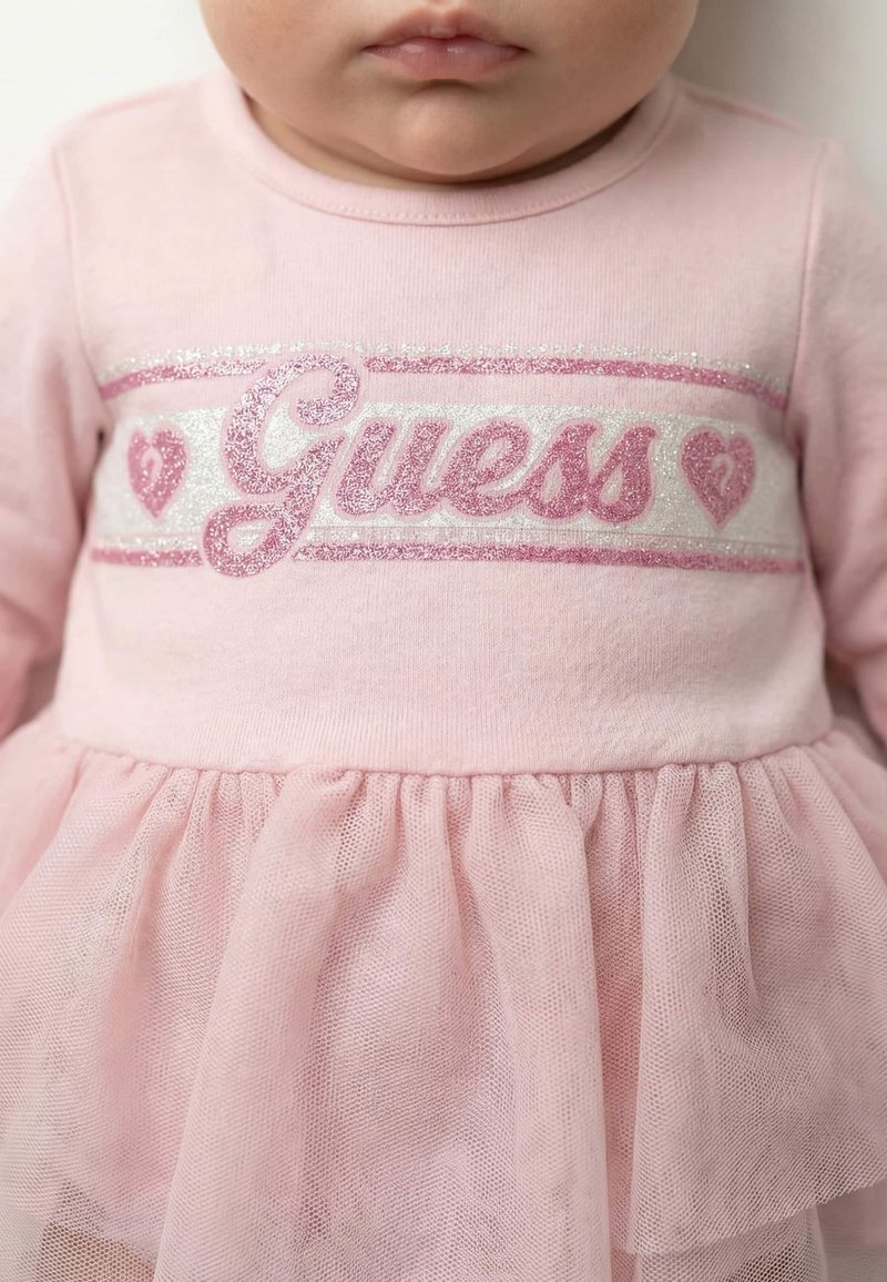 Bébé portant une robe rose avec un texte scintillant "Guess" et des motifs en forme de cœur sur la poitrine, avec une jupe en tulle.