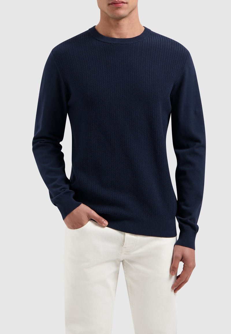 Marineblauer Strickpullover mit strukturiertem Muster, Rundhalsausschnitt und langen Ärmeln, kombiniert mit hellen Hosen und Händen in den Taschen.