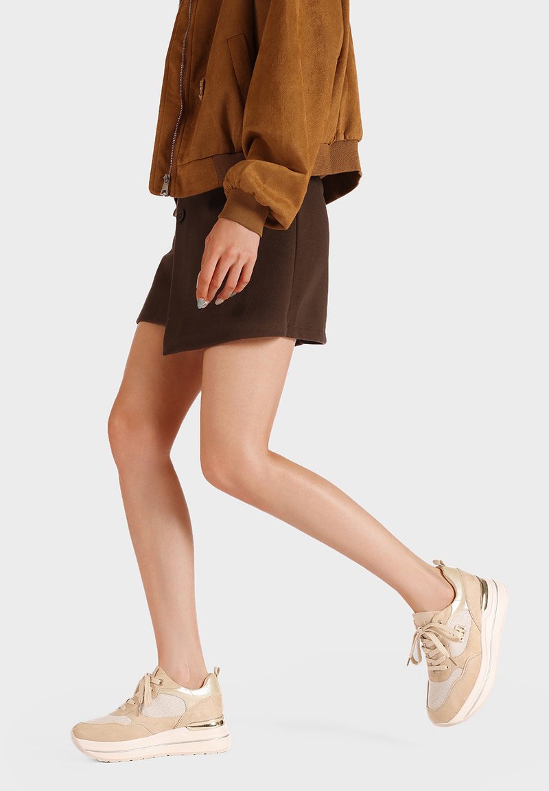 Scarpe sportive beige con una combinazione di suede e rete, caratterizzate da dettagli metallici, suola imbottita e design con lacci, indossate con una gonna marrone.