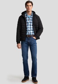 Giacca nera imbottita con cappuccio sopra una camicia a quadri blu e nera, abbinata a jeans blu e sneaker scure. Design minimalista.