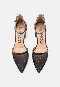 Svarte sko med hæl og spiss tå, overdel i gjennomsiktig mesh med hvite polkaprikker, og ankelrem. Merkevarenavnet er synlig på innersålen.