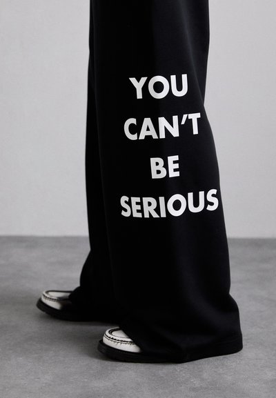 Čierne tepláky s výrazným bielym nápisom "NEMÔŽEŠ TO MYSLIEŤ VAŽNE." Oblečenie je doplnené o biele topánky s kontrastnou čiernou podrážkou.