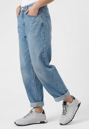 Osoba nosící zastrčené světle modré džíny a bílé tenisky s černými detaily, stojící s rukama v kapsách před jednobarevným pozadím.