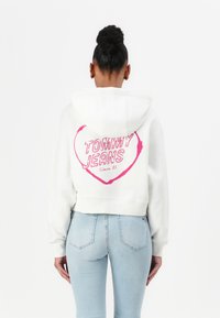 Haut à capuche blanc raccourci avec un graphique de cœur rose portant l'inscription "TOMMY JEANS Depuis 85" au dos. Assorti à un jean ajusté bleu clair.