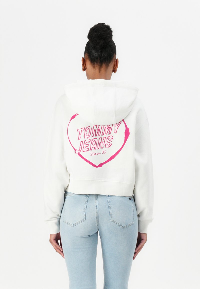 Haut à capuche blanc raccourci avec un graphique de cœur rose portant l'inscription "TOMMY JEANS Depuis 85" au dos. Assorti à un jean ajusté bleu clair.