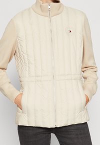 Beżowa pikowana kamizelka z wysokim kołnierzem, przednim zamkiem błyskawicznym i dwoma bocznymi kieszeniami. Na piersi znajduje się naszywka z logo, a mankiety są ściągane.