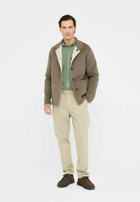 Chaqueta de mezcla de lana marrón con un cuello blanco, camisa verde de botones, pantalones beiges y zapatos marrones. Pose activa con las manos en los bolsillos.