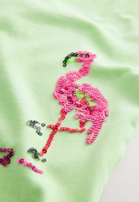 Vihreä kangasvaate, jossa on vaaleanpunaisilla paljeteilla varustettu flamingokuvio, mustilla ja vihreillä yksityiskohdilla, mikä tuo esiin teksturoidun, kiiltävän pinnan.