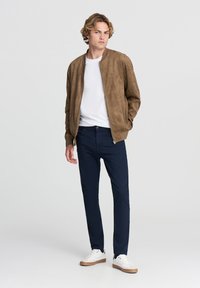 Brązowa skórzana kurtka bomberka noszona na białym podkoszulku, połączona z granatowymi spodniami i białymi sneakersami. Kurtka ma zamek błyskawiczny z przodu oraz ściągacze na mankietach.