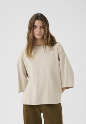 Beige oversized shirt with short sleeves, featuring a round neckline and subtle seam detailing. Fabric appears soft and smooth.