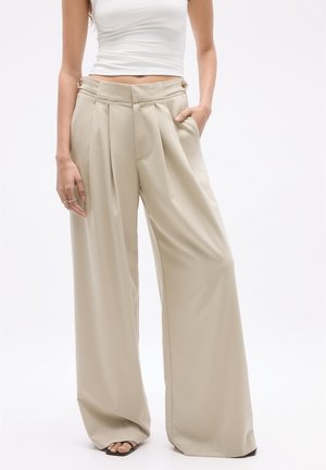 Woman wearing high-waist beige wide-leg trousers with pleats, buttons at waist, white cropped tank top, and black open-toe sandals.
